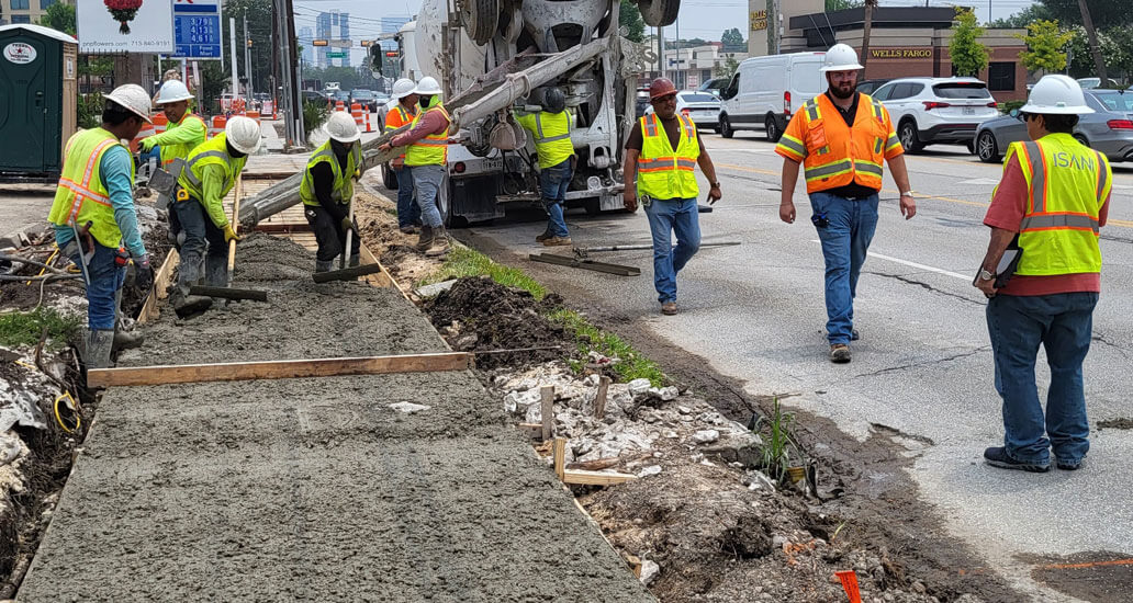 Truck pours concrete while workers spread it out a on the sidewalk.